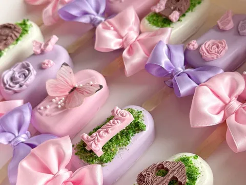 A dozen pastel cakesicles in purple, pink, and white, decorated with satin bows, butterflies, roses, and a number-one topper.