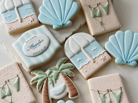 Chic Set sugar cookies with a coastal beach-club theme: striped "San Diego Beach Club" plaques, palm trees, seashells, and bikinis in blue and sand tones.