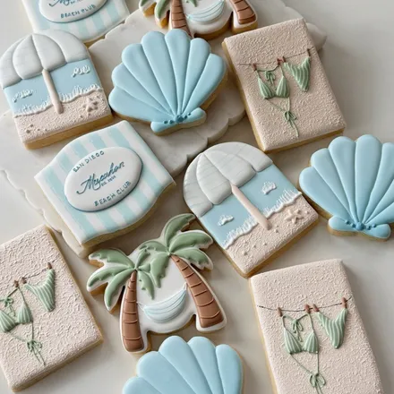 Chic Set sugar cookies with a coastal beach-club theme: striped "San Diego Beach Club" plaques, palm trees, seashells, and bikinis in blue and sand tones.