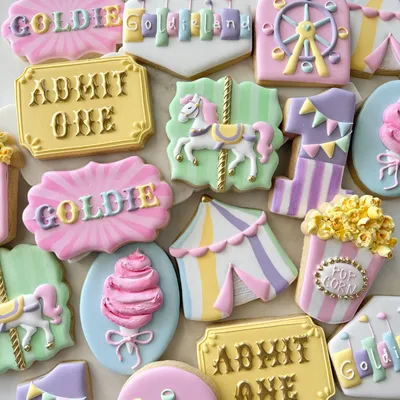 Pastel circus-themed birthday cookie set for "Goldie" with a carousel horse, ferris wheel, popcorn box, circus tent, and cotton candy in pink, lavender, and yellow.