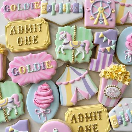 Pastel circus-themed birthday cookie set for "Goldie" with a carousel horse, ferris wheel, popcorn box, circus tent, and cotton candy in pink, lavender, and yellow.