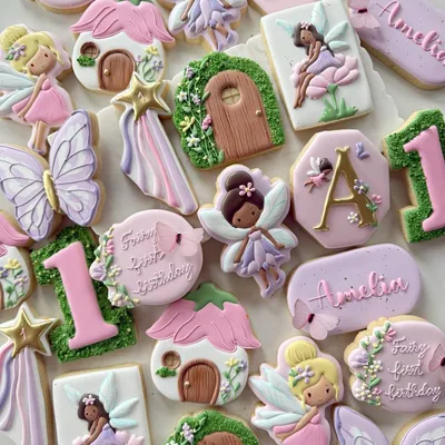 Pastel fairy-themed first birthday cookie set for "Amelia" with fairy characters, a flower cottage door, butterflies, and a shooting-star wand in pink, lavender, and green.