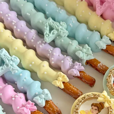 Caramel and chocolate-dipped pretzel rods in a ridged rainbow of pastel mint, blue, lavender, yellow, and pink, finished with bows and a number topper.