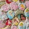 Disney princess-themed 4th birthday cookie set for "Princess Sophia" with hand-painted portraits of Ariel, Belle, Moana, Elsa, Aurora, and Cinderella, a castle, tiara, and pumpkin carriage in pink and gold.
