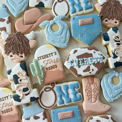 Western rodeo-themed first birthday cookie set for "Everett" with a cow-print pattern, cowboy boot, bandana, lasso, cactus, and highland cattle in blue, brown, and cream.