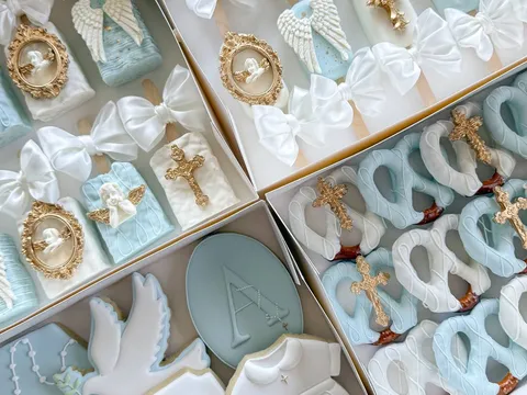 Small party package with christening-themed treats: angel-wing and gold-cross cake pops, a dove cookie, and a "God Bless Asher" plaque in blue, white, and gold.